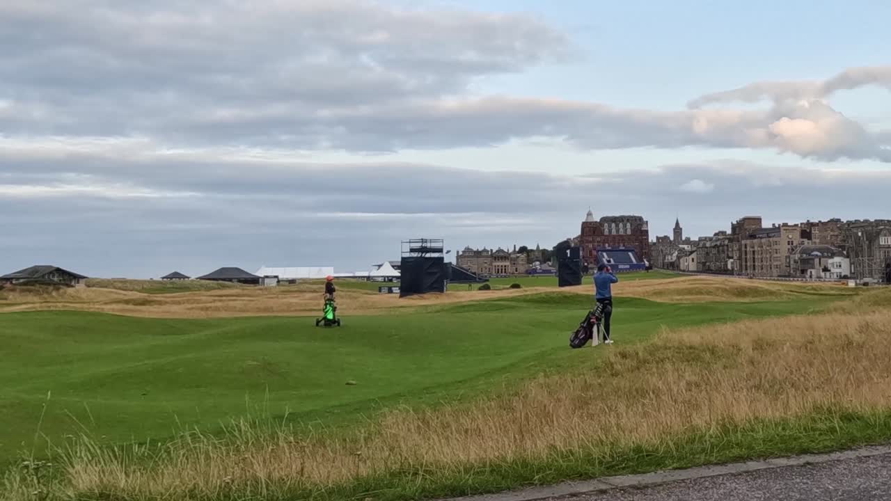 Golfers walk across a vibrant green fairway beneath a cloudy sky, with distant buildings in view.