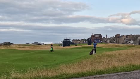 golfers walk across a vibrant green fairway beneath a cloudy sky, with distant buildings in view.