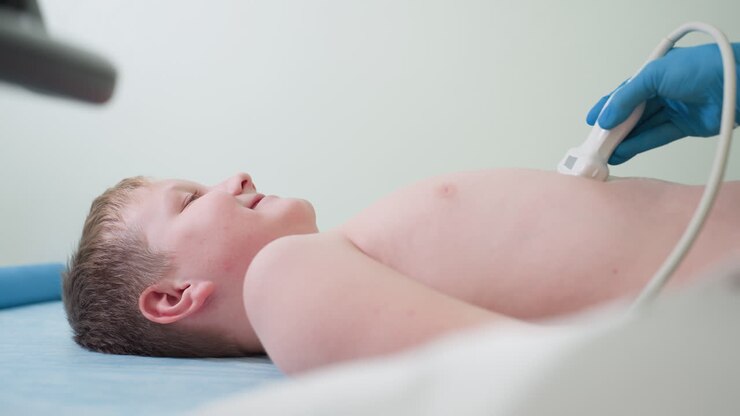 Young boy lies comfortably on clinical bed smiling while being examined with diagnostic transducer by medical professional, showing calmness and ease during procedure in clean healthcare environment