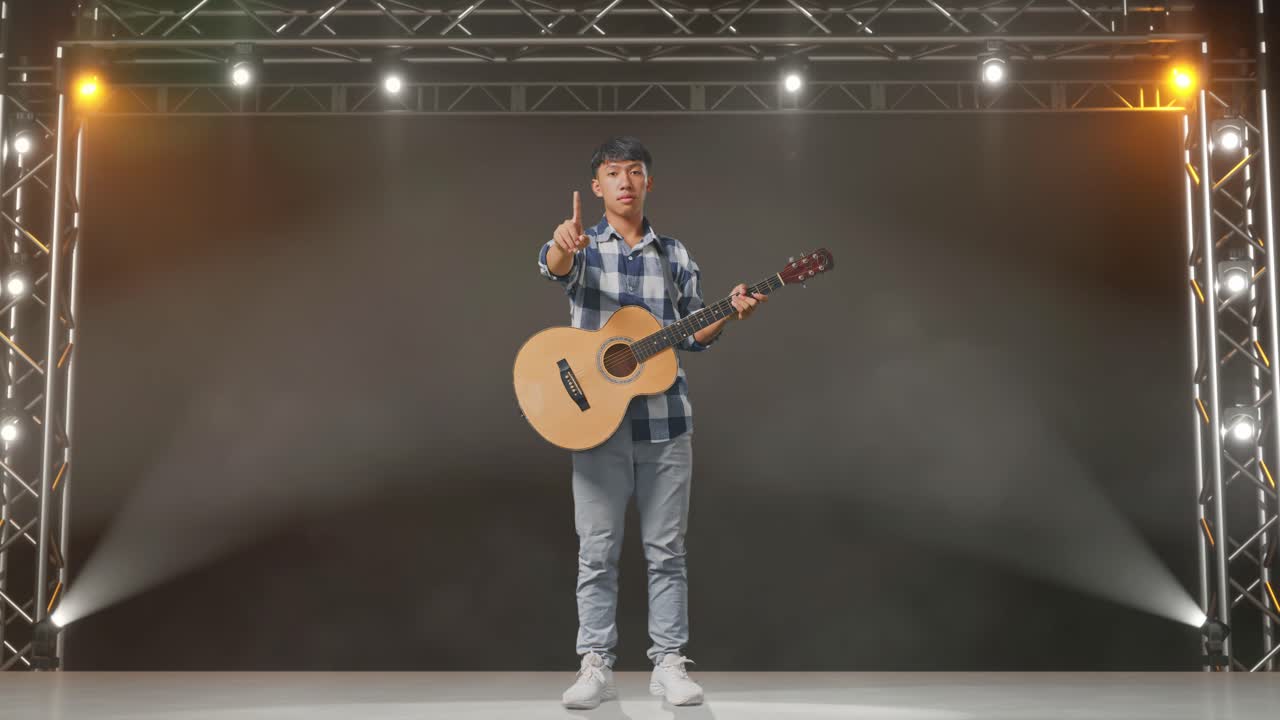 cuerpo lleno de adolescente asiático con guitarra que no muestra ningún gesto mientras está de pie en el escenario