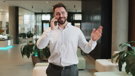 un joven empresario caucásico sonriente hablando por teléfono inteligente discutiendo el trabajo en el vestíbulo de la oficina del hotel