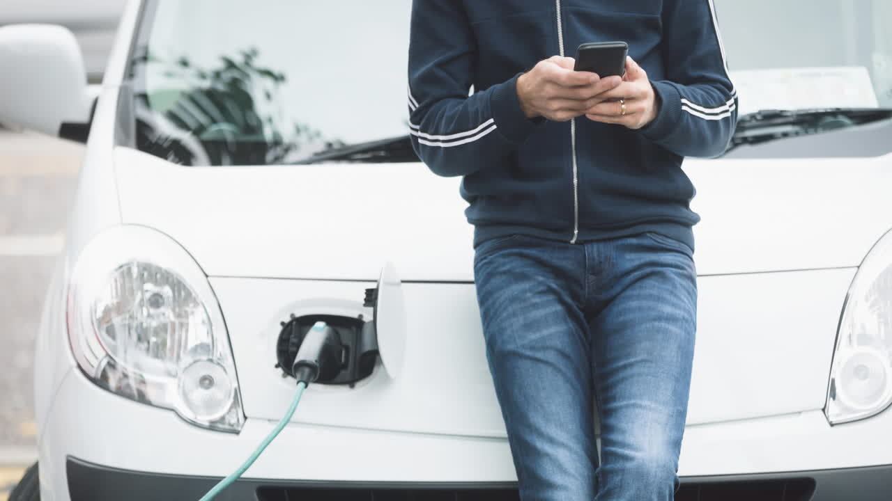 sección media de hombre caucásico usando un teléfono inteligente y cargando un coche eléctrico