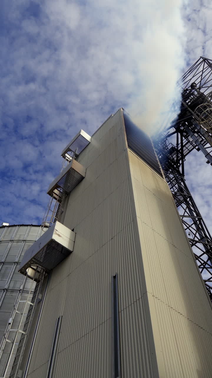 Grain elevators against blue sky. Thick white smoke releases from industrial building. Modern complex for agribusiness. View from below. Vertical video