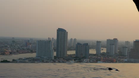 Person,-Die-In-Einem-Infinity-Pool-Auf-Einem-Hochhaus-In-Bangkok-Schwimmt,-Mit-Flussblick-Auf-Die-Skyline-In-Thailand