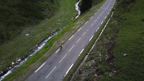 Drohnen-Luftschwenkansicht-Eines-Radfahrers,-Der-Eine-Friedliche-Straße-In-Den-Französischen-Pyrenäen-Neben-Einem-Kleinen-Fluss-Mit-Grünem-Gras-In-Den-Bergen-Hinaufklettert