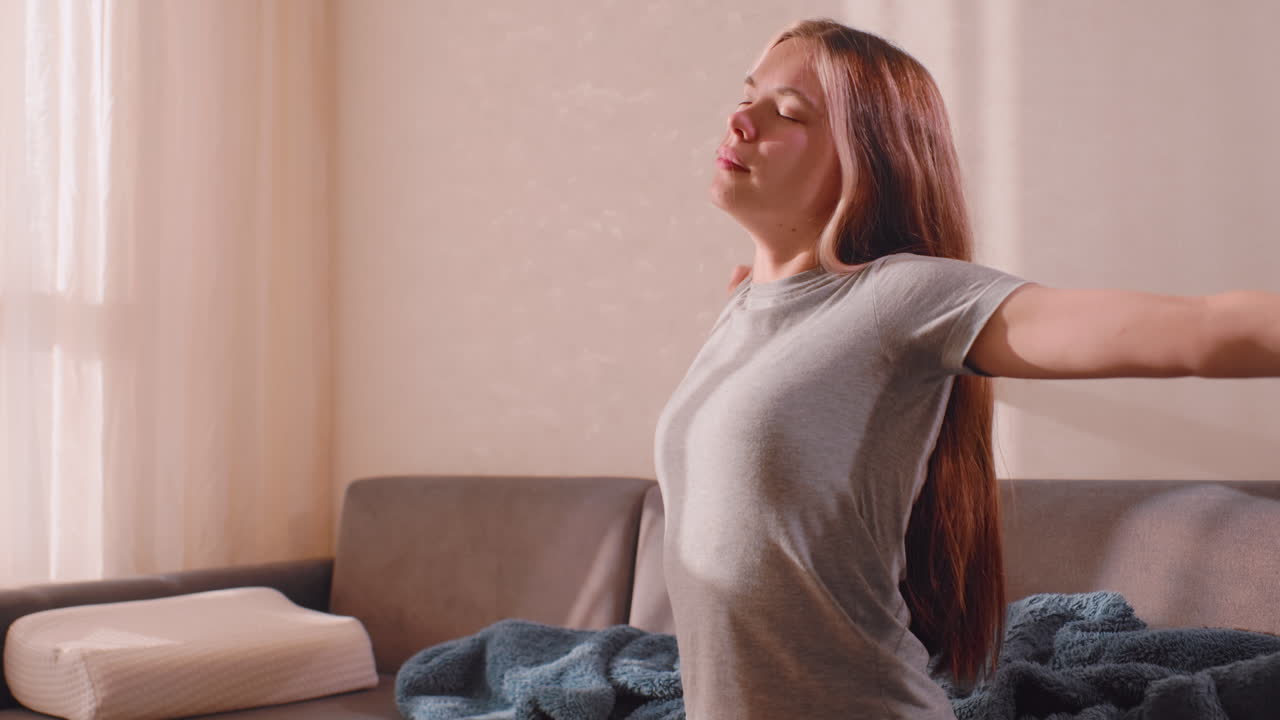 Teenage girl with long hair sits on sofa near window stretching arms gently after waking, eyes closed, soaking in early sunlight through sheer curtains in quiet cozy morning