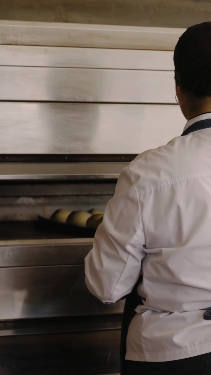 Video verticale: Forno a piani che emette un segnale acustico, fornaio con giacca da chef e grembiule scuro che estrae un vassoio in panetteria.