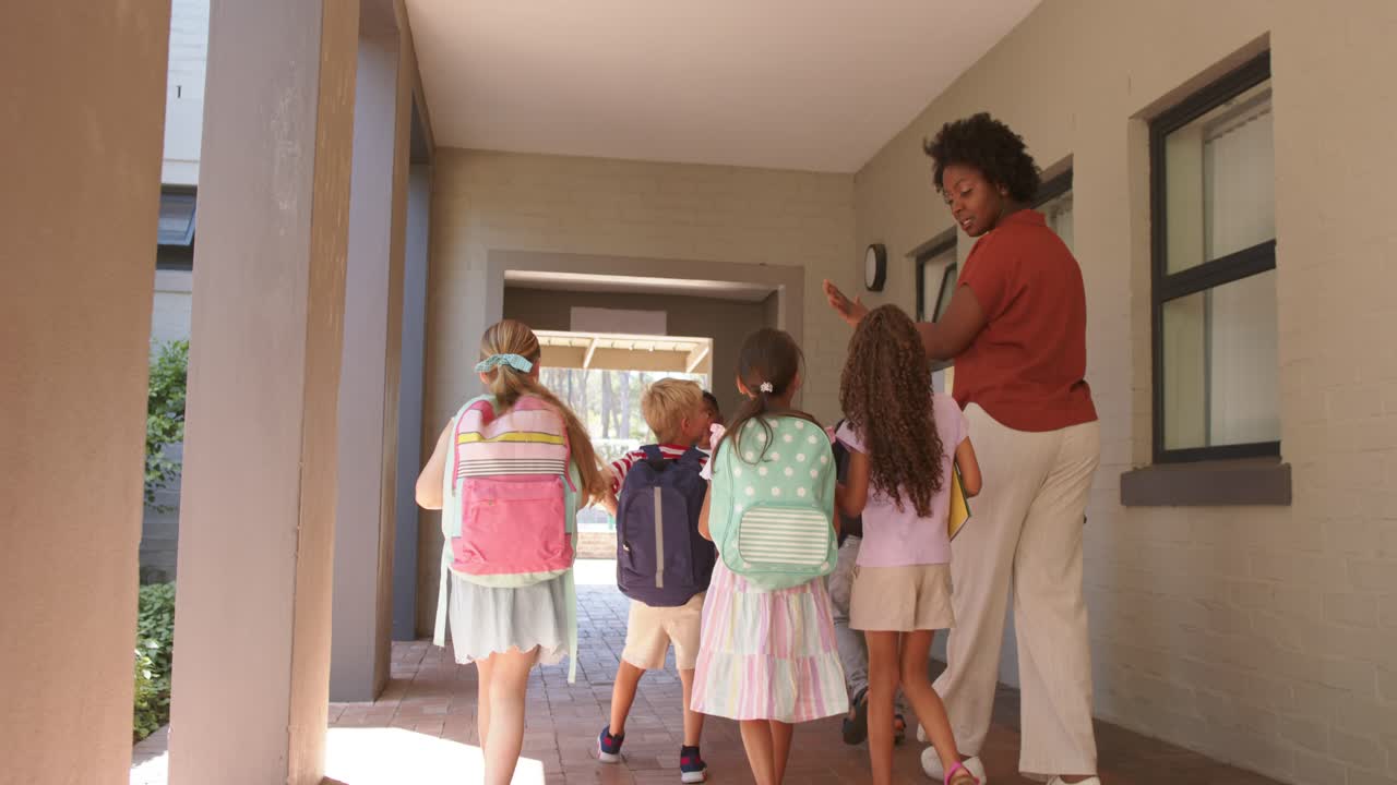 African American woman raising hand with tablet leading kids with backpacks down hall to courtyard