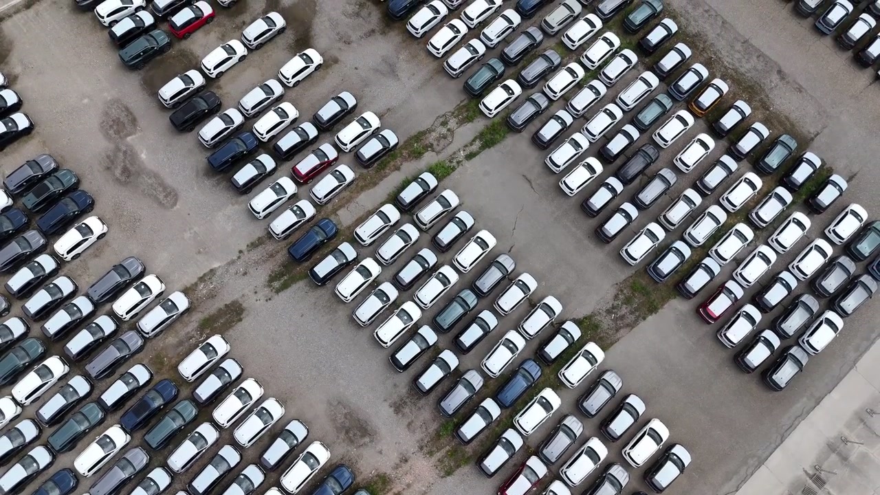 Personbiler er arrangeret i tætte opbevaringsrækker inde i et stort logistikområde for biler til flådedistribution, opadstigende droneoptagelse fra oven