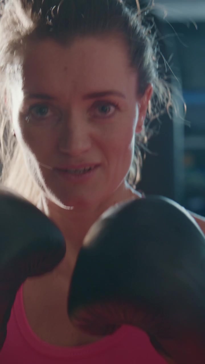 Una pugile concentrata si allena in una palestra poco illuminata. Il suo sguardo è intenso, i muscoli tesi. Indossa abbigliamento sportivo rosa e guantoni neri. Si muove con determinazione, le ombre danzano sul suo viso.