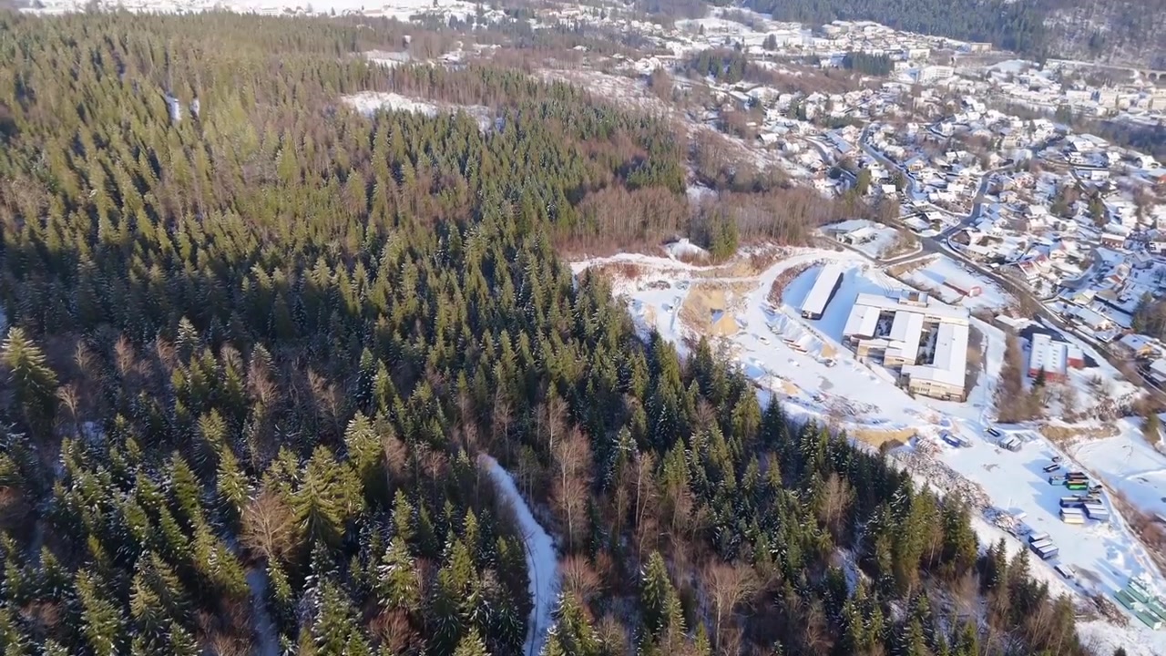 Vozidlo na solární pohon zaparkované v zasněženém lese, zatímco dron se zvedá ze strmého pohledu shora dolů, odhalující jehličnatou lesní krajinu v Regionálním přírodním parku Haut-Jura ve Francii.
