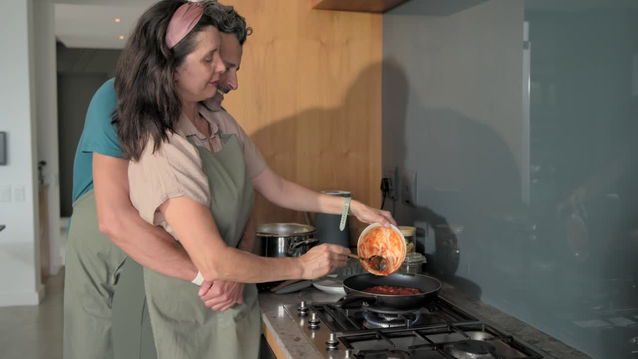 Coppia che cucina, donna che versa salsa di pomodoro in padella su un fornello acceso mentre l'uomo trita ingredienti per la cena.