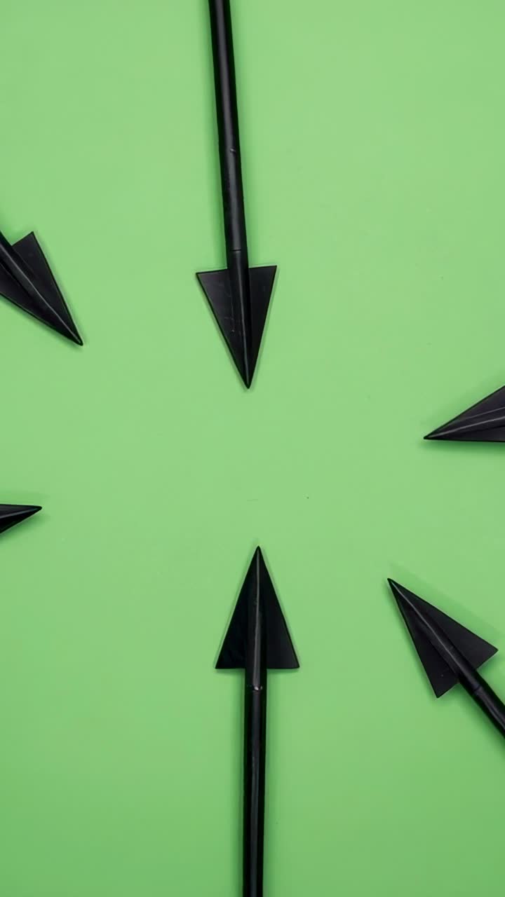 Vertical video: Four black metal arrowheads pointing inward on light green surface, directing focus
