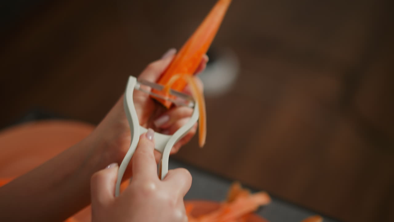 Close up of person holding pine-shaped carrot with white peeler in hand, blur background with indistinct object, focus on orange vegetable and kitchen tool during food preparation process