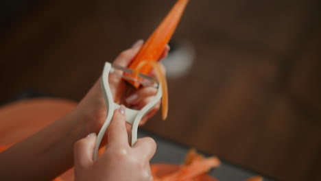 close up of person holding pine-shaped carrot with white peeler in hand, blur background with indistinct object, focus on orange vegetable and kitchen tool during food preparation process