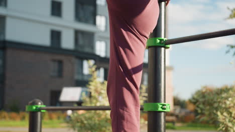 back view of motivated girl holding bar and extending side leg high while standing on other foot under bright sun in urban park workout in hoodie vest showcasing strength flexibility focus outdoors