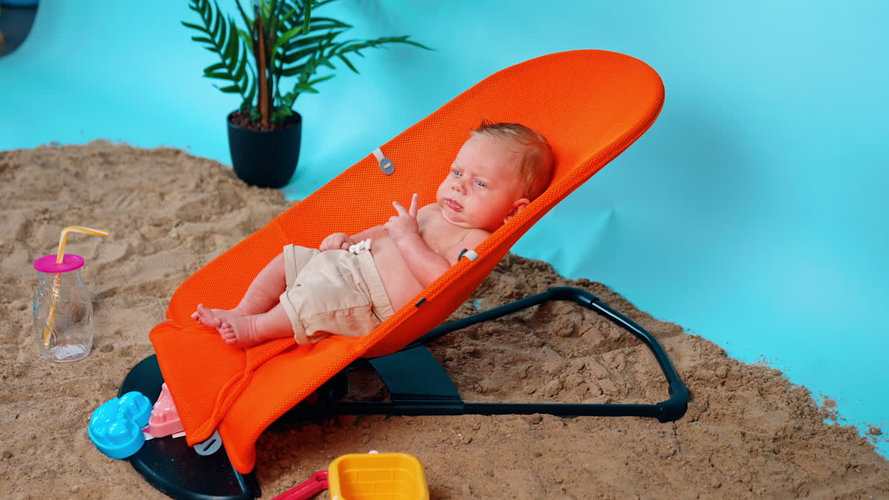 Serious newborn boy in shorts sitting in the orange baby chair. Kid resting on the improvised beach in studio.