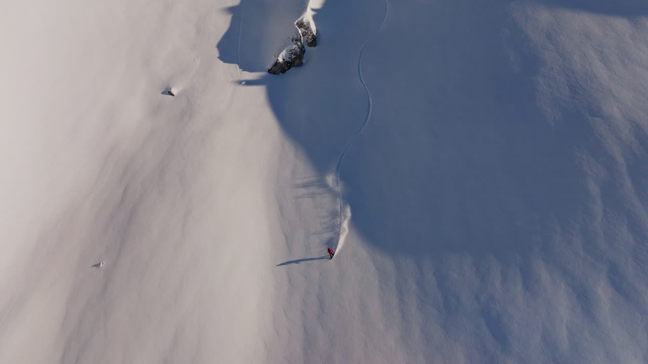 Aerial drone shot of a snowboarder carving fresh powder down steep Verbier slopes on a pristine bluebird day, white spray and dramatic Alpine peaks in the background