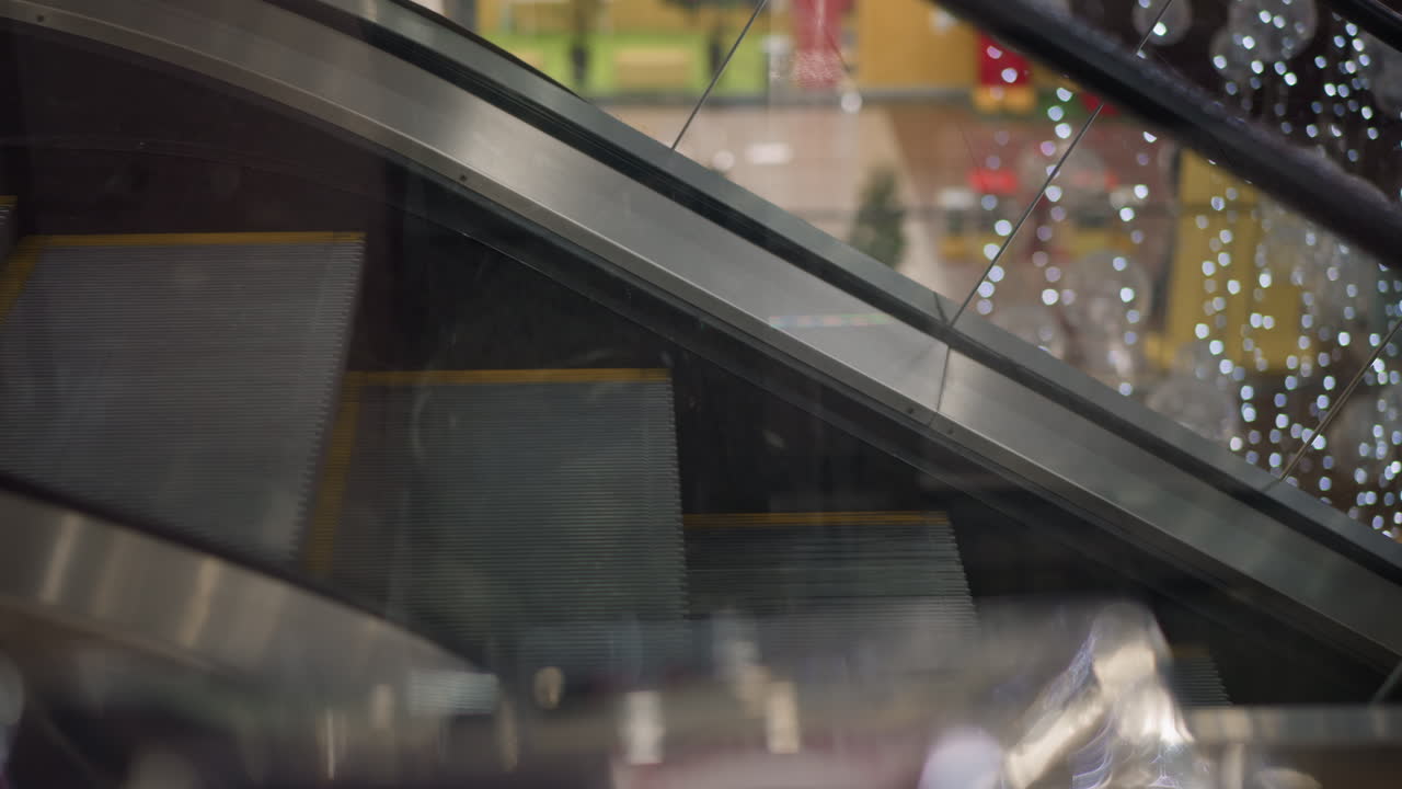 glass panel reflection showing escalator steps in motion beneath decorative hanging lights inside modern mall interior captured through transparent barrier reflections of lights shadows shoppers