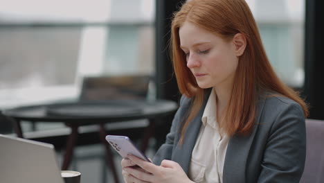 pretty redhead woman in grey jacket is working in co-working area viewing social media and message in smartphone