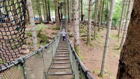 cute young blonde girl waking on rope suspension bridge inside woods of mikkelparken theme park - norway