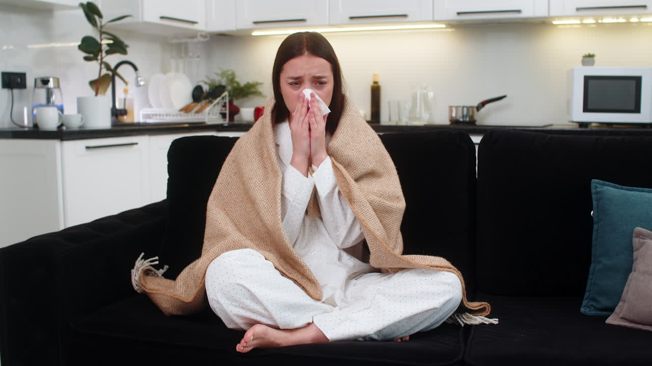 Giovane donna avvolta in una coperta starnutisce, usa un fazzoletto, ha sintomi di raffreddore e influenza, tira su col naso, non sta bene, è malata, stanca e debole