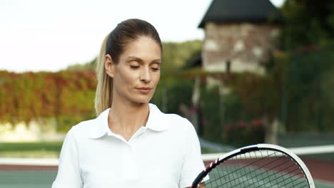 Porträt-Einer-Schönen-Frau-Im-Weißen-Poloshirt,-Die-Mit-Schläger-über-Der-Schulter-Auf-Einem-Tennisplatz-Steht-Und-Freudig-In-Die-Kamera-Lächelt