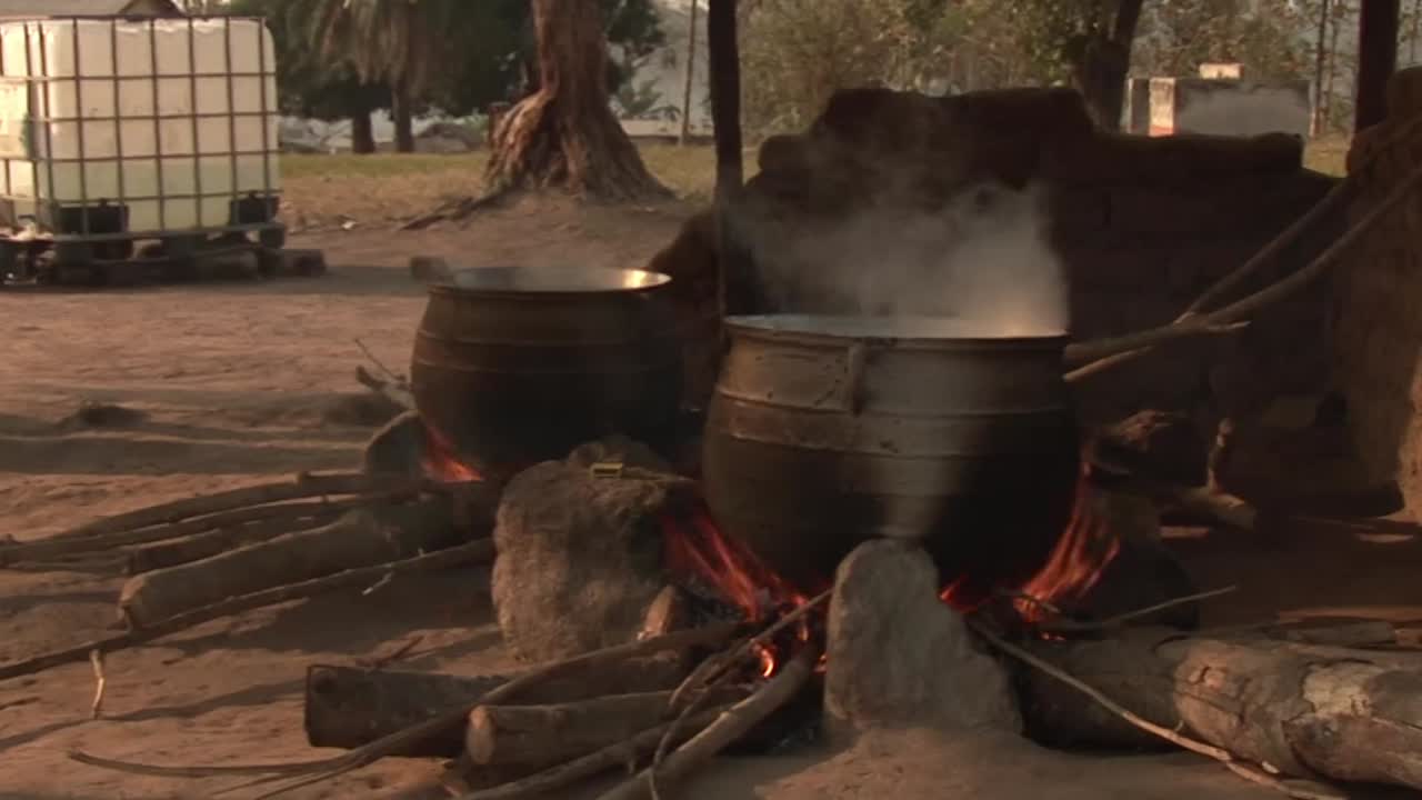 Large metal pots boiling over open wood fires in outdoor kitchen, steam rising as food cooks in rural West African setting