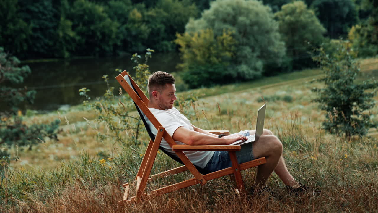 Male freelancer sits in a chair with laptop on his knees. Side view. Beautiful nature around.