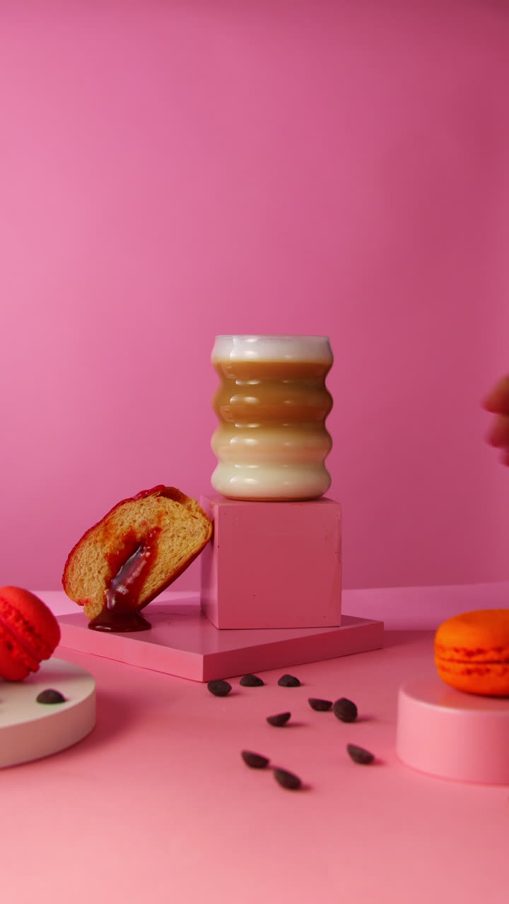 Layered cappuccino is placed next to a sliced jelly donut and chocolate chips on a pastel pink stage, perfect for café branding, online marketing, or dessert product promotion
