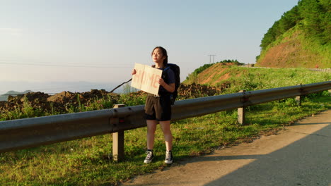 Mujer-Asiática-Excursionista-Con-Mochila-Sostiene-Un-Cartel-De-Cartón-Con-La-Casa-Escrita-En-él,-Autoestopista-Al-Costado-De-La-Carretera-En-Asia