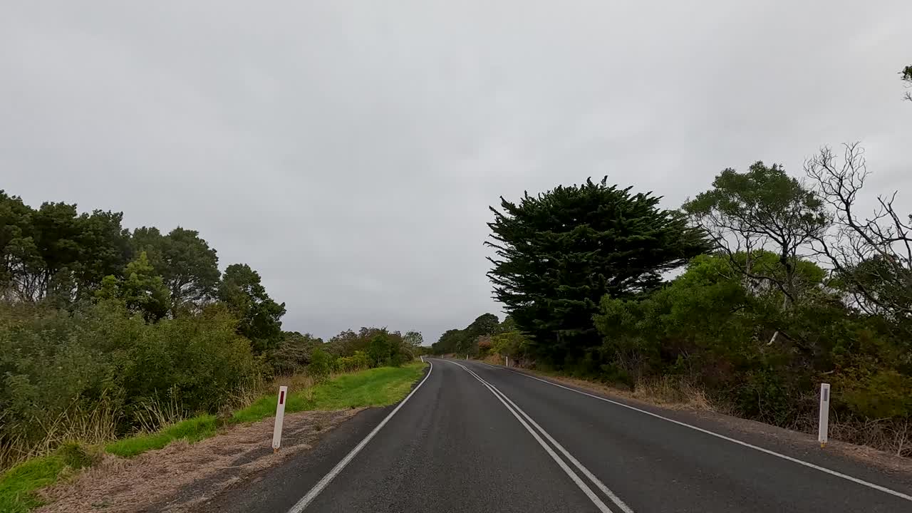 A fast-motion sequence traveling down a two-lane asphalt road through open countryside, featuring scattered trees, grassy fields, and overcast daylight with smooth forward camera movement