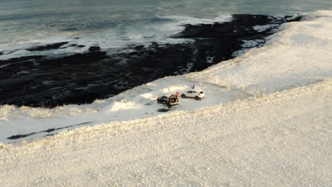 drone orbits around cars parked at top of snow covered sea cliff