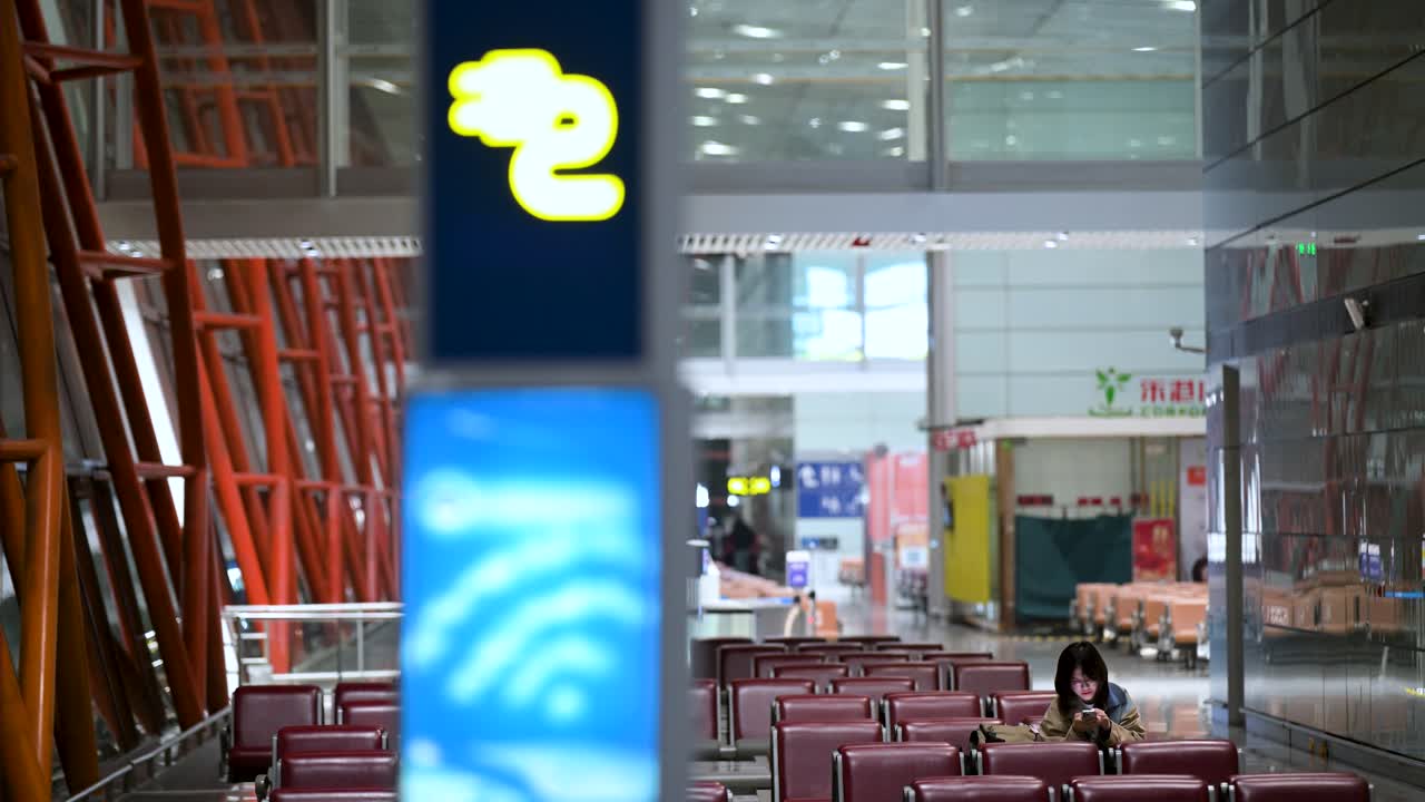 A charging station sign is seen in the foreground as a female flight passenger uses a smartphone while sitting and waiting at the boarding gate in Beijing International Airport, China.