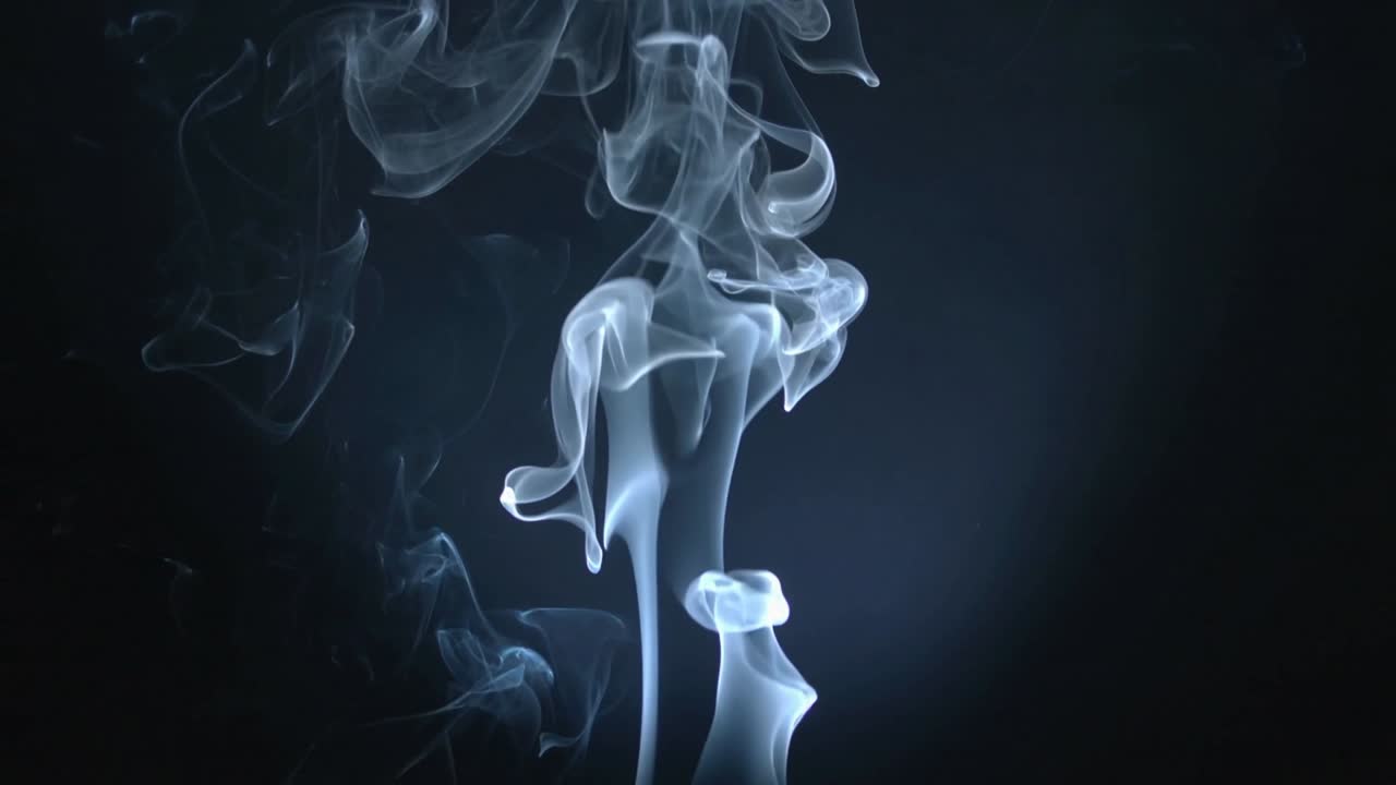 Colonna di fumo verticale che sale, rispondendo a un soffio iniziale, formando spirali in studio con una retroilluminazione fredda.