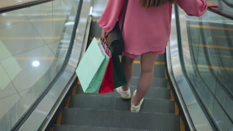 back view of lady in pink dress holding colorful shopping bags and black handbag while standing on escalator, she rides downwards in a modern mall, light is seen reflecting on the glass