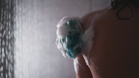 woman rubs skin with loofah in shower cabin closeup. lady washes body with soft foamy sponge near falling water drops. skincare and massage tool