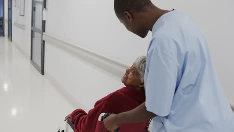 diverse female doctor pushing senior female patient in wheelchair in hospital corridor, slow motion