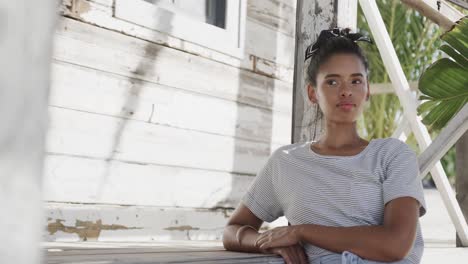 thoughtful biracial woman standing outside wooden beach house, in slow motion