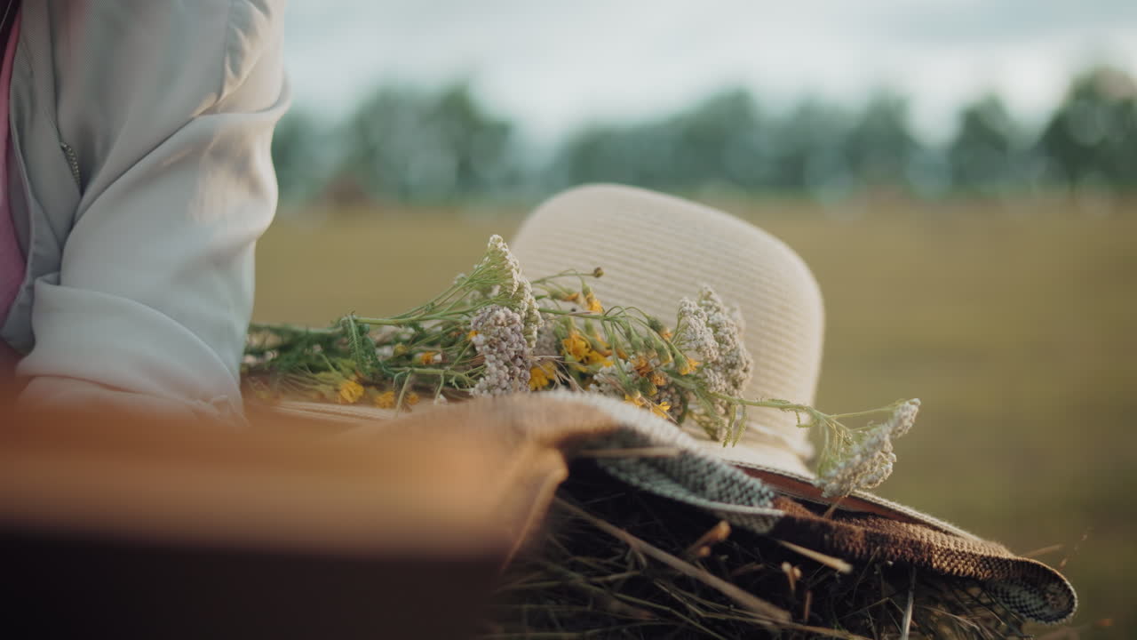 Close-up of a sun hat and wildflowers resting on a blanket over a hay bale, with a soft-focus background of an open field and a partial view of a person