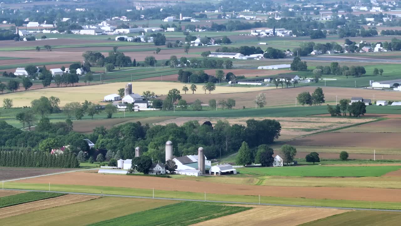 Expansive view of the American countryside with vast fields, scattered farmhouses and silos under clear sky. Landscape captures the essence of rural life and agricultural abundance. Aerial wide shot.