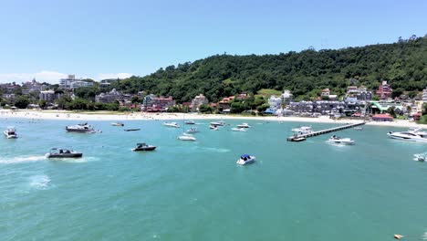 escena aérea de drones de playa turística en florianópolis brasil con muchos botes y lanchas rápidas en el mar con muelle y mucha gente navegando junto a la playa con muchas casas frente al mar