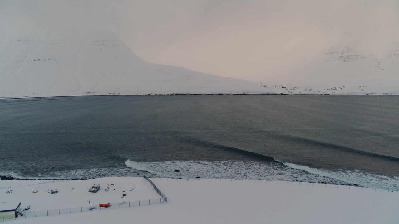 Aerial tracking above ocean waves crashing on a vast snowy Arctic landscape with gentle dusk lighting in Iceland