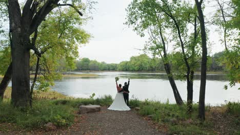 aerial, newly wed heterosexual couple raising arms up celebrating marriage in outdoor forest by a river