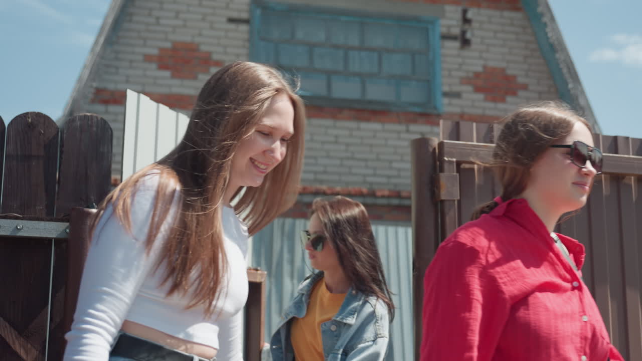 Happy lady opens door for two friends while last friend closes it behind her, stepping onto paved yard with green grass beside wooden fence under clear blue sky in suburban setting