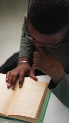 black man reads book sitting at table upper view. student studies textbook doing graduate work in reading hall. african american guy in library