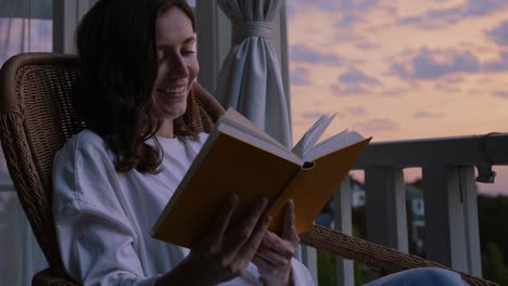 relaxed woman enjoying reading book while sitting on rocking chair at home balcony during vibrant sunset with colorful sky glowing softly