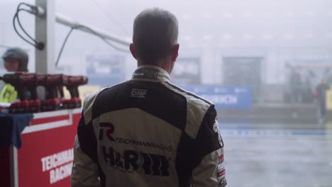 race car driver in fireproof suit looking at foggy wet pitlane bad weather conditions from inside box paddock garage at the adac nurburgring twentyfour hour race.