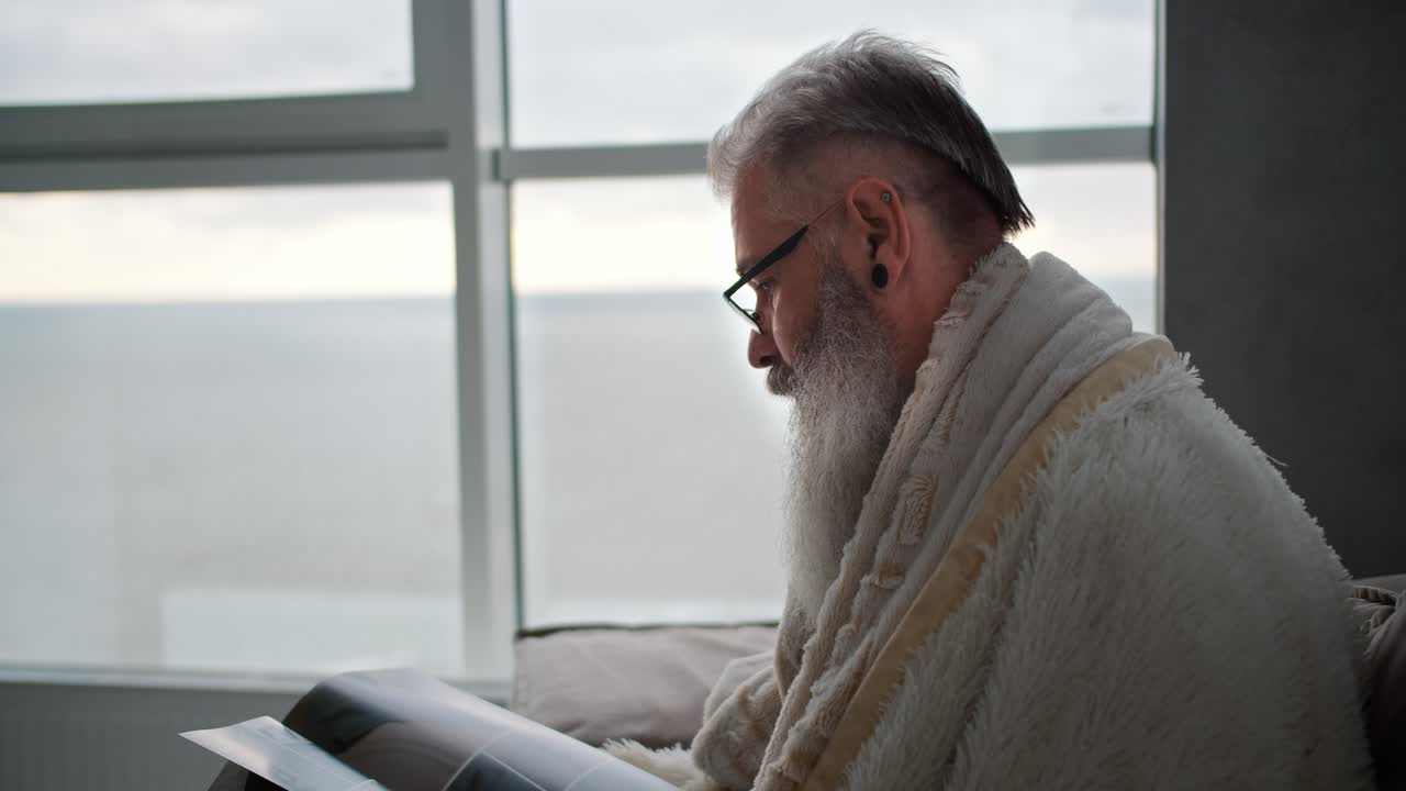 An elderly man with gray hair and a lush beard is wrapped in a white blanket looking at a magazine while sitting on a brown sofa in a modern apartment overlooking the sea