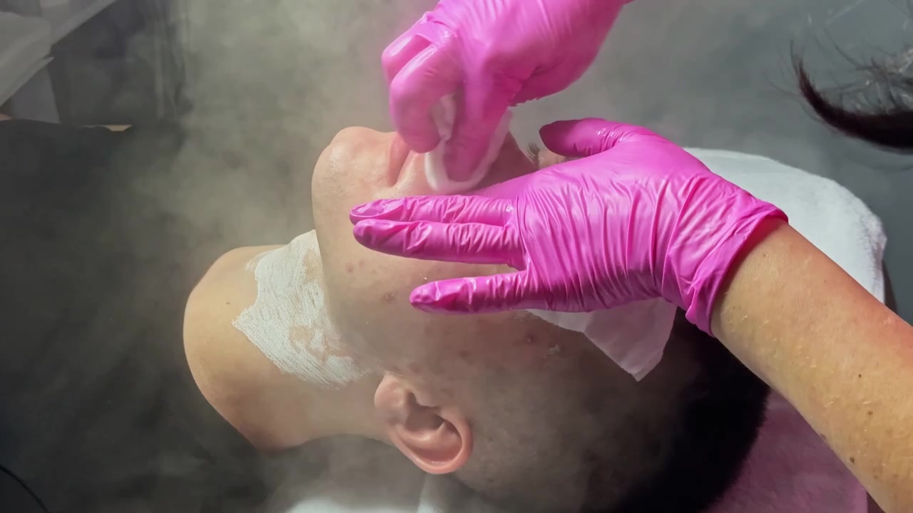 Hombre recibe tratamiento facial en un spa mientras sube vapor y un profesional aplica productos durante una sesión de cuidado de la piel.